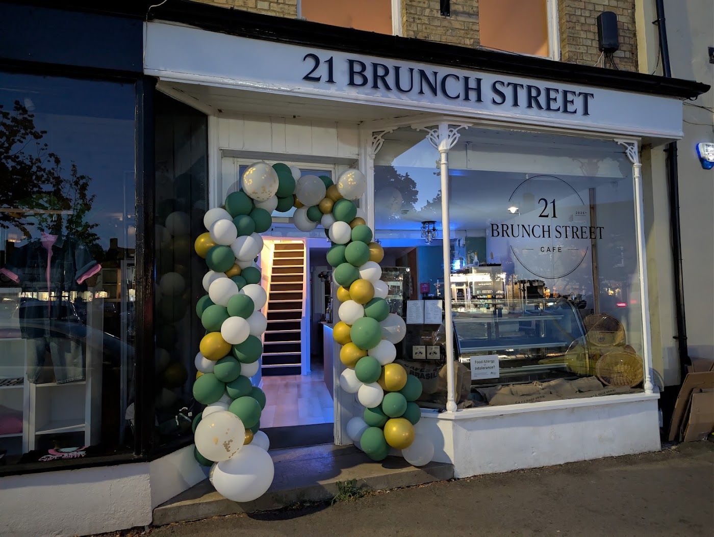 The 21 Brunch Street shopfront on Market Place: a white fascia lettered 21 BRUNCH STREET in black serif capitals, the badge on the window, the cake counter inside and the stairs up to the seating, with a green, white and gold balloon arch round the door